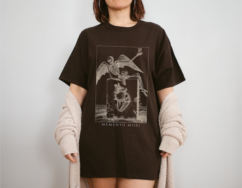 May include: A brown t-shirt with a graphic print of a skeleton with wings sitting on a heart. The text "MEMENTO MORI" is printed below the graphic.