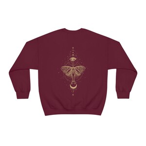 Puede incluir: Una sudadera color burdeos con un gr&aacute;fico dorado de una polilla con un dise&ntilde;o de ojo y luna.