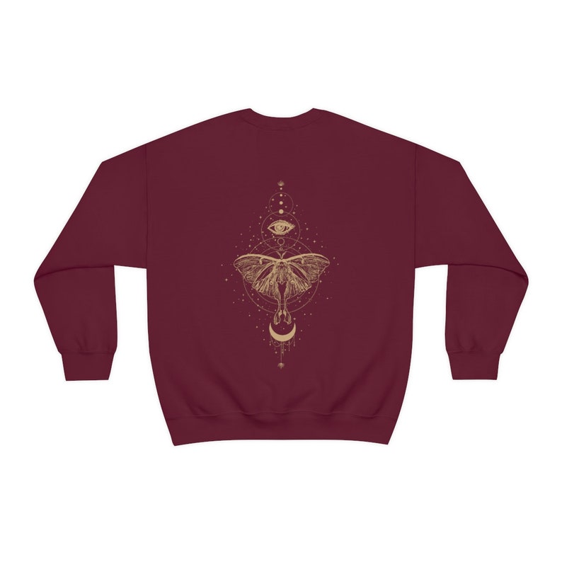 Puede incluir: Una sudadera color burdeos con un gr&aacute;fico dorado de una polilla con un dise&ntilde;o de ojo y luna.