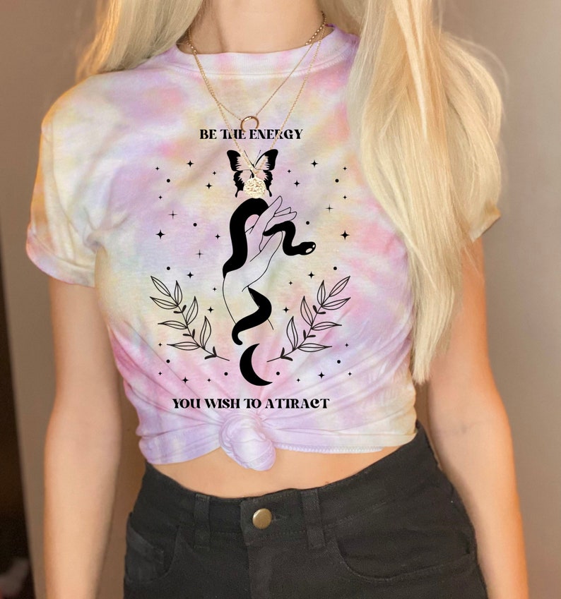 以下が含まれることがあります： パステルカラーのタイダイTシャツ。黒と白のグラフィックで、手に蛇が巻き付き、その頭に蝶がとまっているデザイン。Tシャツには「BE THE ENERGY YOU WISH TO ATTRACT」という文字がプリントされている。