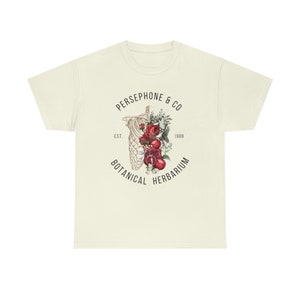 May include: A beige t-shirt with a black and white graphic design of a skeleton with red roses and pomegranates. The text "PERSEPHONE & CO" is above the graphic, and "EST. 1989 BOTANICAL HERBARIUM" is below the graphic.