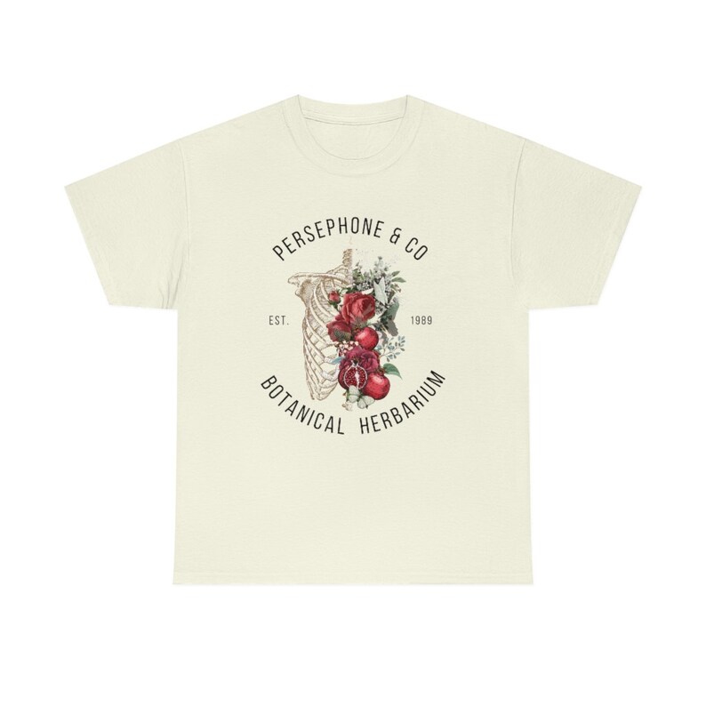 May include: A beige t-shirt with a black and white graphic design of a skeleton with red roses and pomegranates. The text "PERSEPHONE & CO" is above the graphic, and "EST. 1989 BOTANICAL HERBARIUM" is below the graphic.