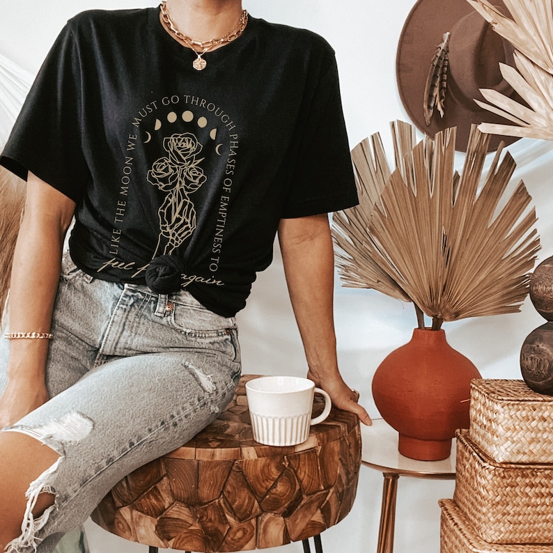 May include: Black t-shirt with a gold graphic of a skeletal hand holding a rose with the text "Like the moon we must go through phases of emptiness to full bloom again".