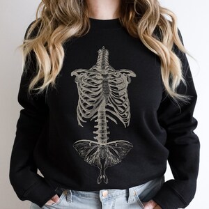 May include: Black sweatshirt featuring a vintage anatomical illustration of a human ribcage and a moth. The moth is centrally positioned and has a detailed design.