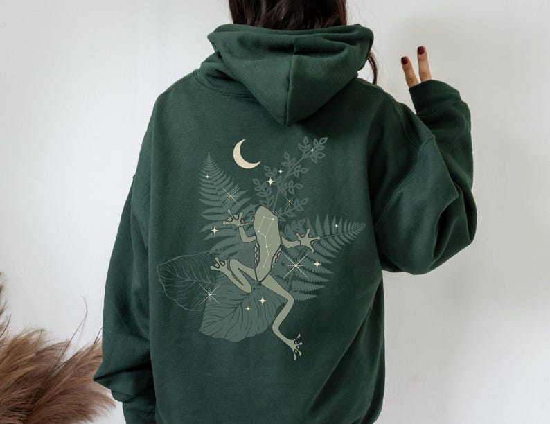 Pu&ograve; includere: Una felpa con cappuccio verde scuro con una stampa grafica di una rana seduta su una felce con una luna crescente sullo sfondo.