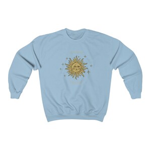 Peut inclure: Sweat-shirt bleu clair avec un motif soleil et &eacute;toiles dor&eacute;s. Le soleil a un visage et est entour&eacute; d'&eacute;toiles. Le texte "The sun always shines" est &eacute;crit autour du soleil.