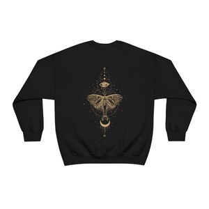 Puede incluir: Sudadera negra con un gr&aacute;fico dorado de una polilla, una luna creciente y un ojo rodeado de estrellas y c&iacute;rculos.