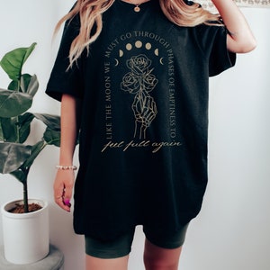 May include: Black oversized t-shirt with a gold graphic design featuring a skeleton hand holding roses and the text "Like the moon we must go through phases of emptiness to get full again."