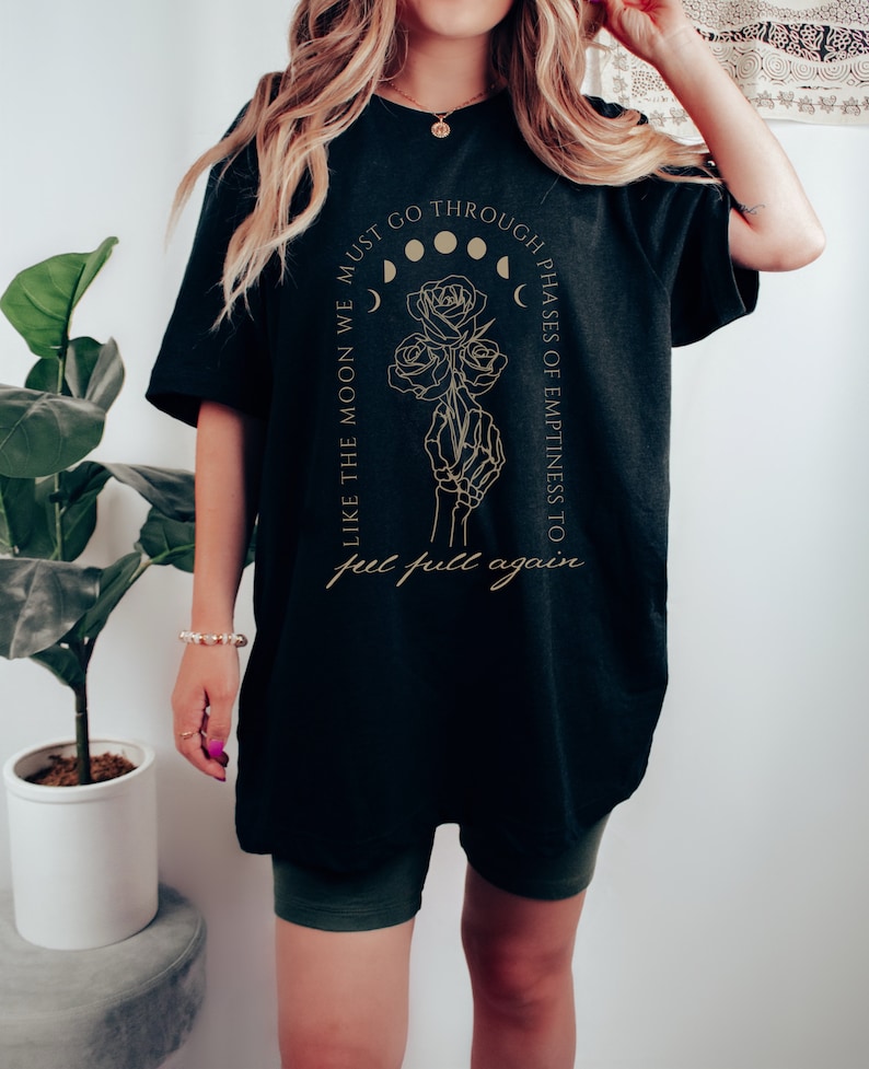 May include: Black oversized t-shirt with a gold graphic design featuring a skeleton hand holding roses and the text "Like the moon we must go through phases of emptiness to get full again."