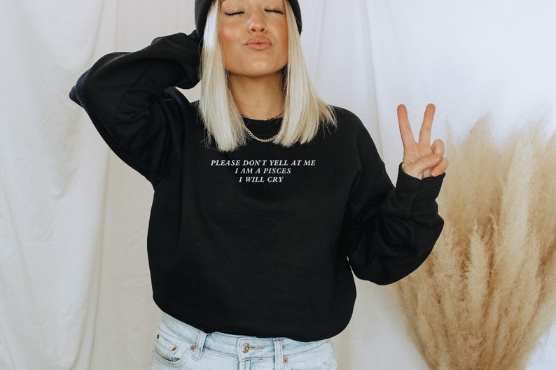 May include: Black sweatshirt with the text "PLEASE DON'T YELL AT ME I AM A PISCES I WILL CRY" in white. The person is wearing a black beanie and light blue jeans. The person is making a peace sign with their fingers.
