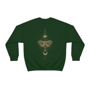 Puede incluir: Una sudadera verde oscuro con un dise&ntilde;o dorado de una polilla con una luna creciente y un ojo en un c&iacute;rculo en la espalda.
