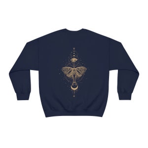 Puede incluir: Sudadera azul marino con un gr&aacute;fico dorado de una polilla, una luna creciente y un ojo rodeado de estrellas y formas geom&eacute;tricas.