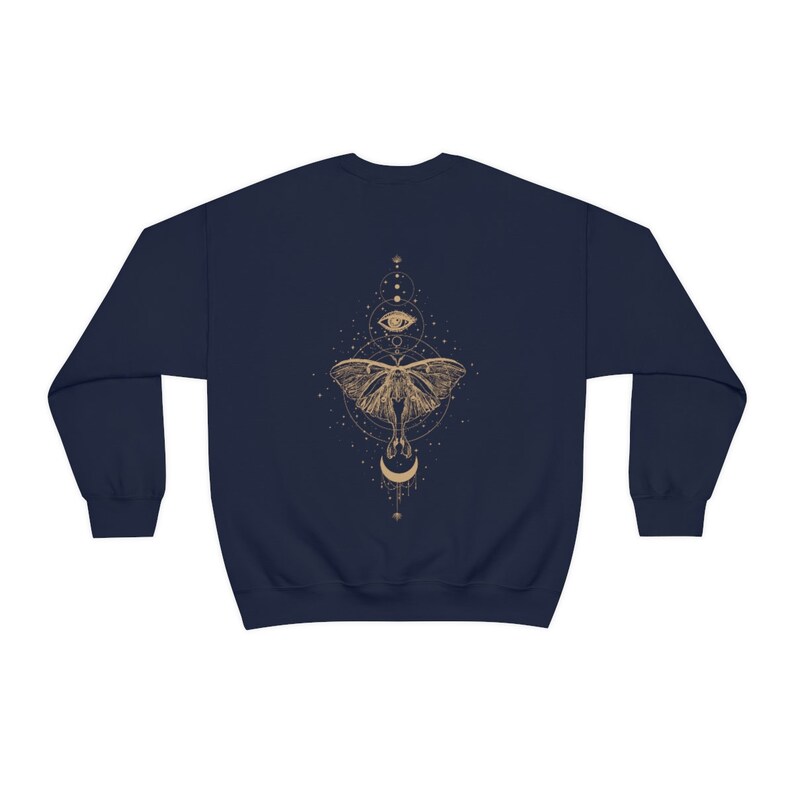 Puede incluir: Sudadera azul marino con un gr&aacute;fico dorado de una polilla, una luna creciente y un ojo rodeado de estrellas y formas geom&eacute;tricas.