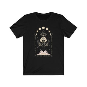 May include: Black t-shirt featuring a mystical design with a woman's face, an eye, and moon phases. The text reads "MAY ALL EVIL EYES IN MY LIFE GO BLIND. PROTECT MY PEACE." The design is in cream and gold tones.