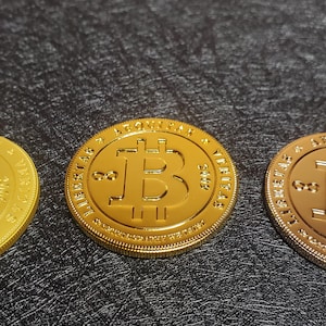Pu&ograve; includere: Tre monete dorate con la scritta "Bitcoin" e il simbolo Bitcoin sul davanti. Le monete sono su una superficie nera testurizzata.