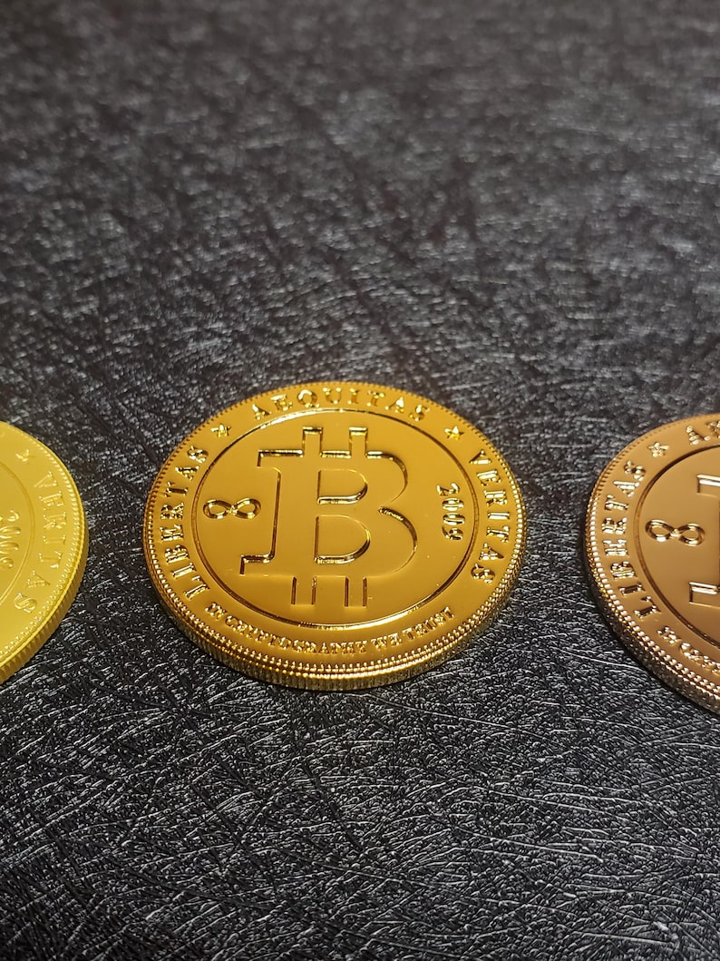 Pu&ograve; includere: Tre monete dorate con la scritta "Bitcoin" e il simbolo Bitcoin sul davanti. Le monete sono su una superficie nera testurizzata.