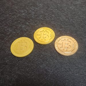Pu&ograve; includere: Tre monete Bitcoin dorate con il simbolo Bitcoin su ciascuna. Le monete sono su una superficie nera.