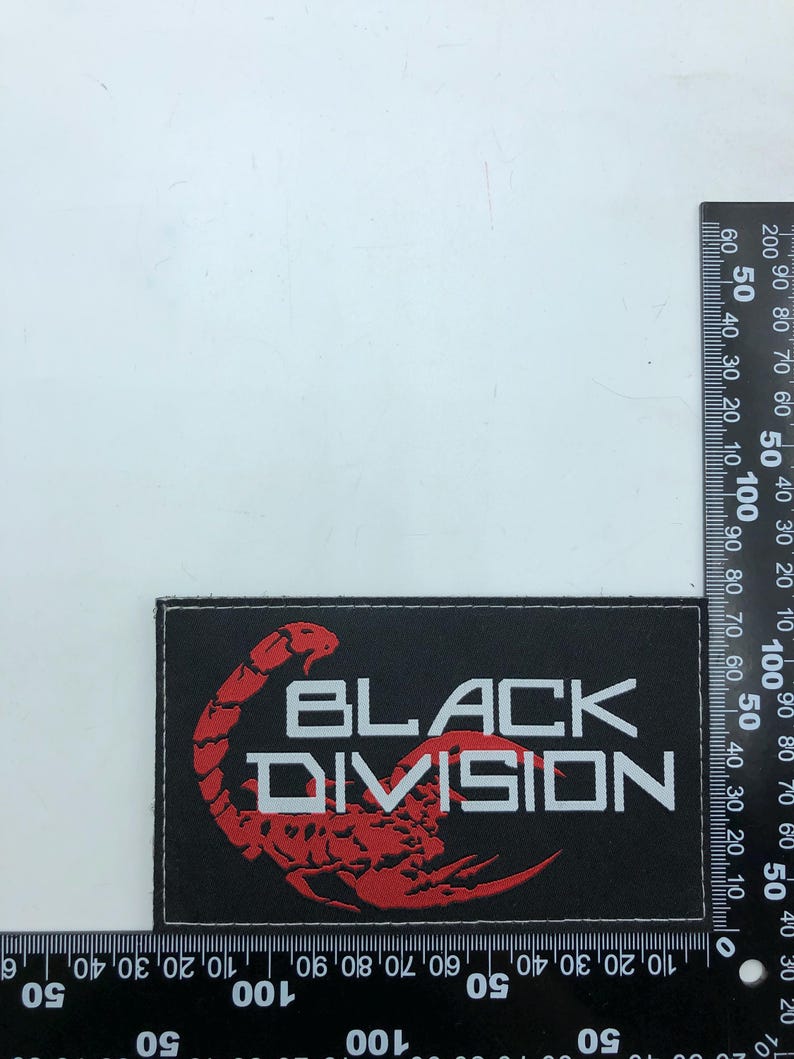 May include: A rectangular black patch featuring a red embroidered scorpion graphic and the white text "BLACK DIVISION". The patch is positioned against a black ruler, showing a width of approximately 9 cm and a height of 5 cm.