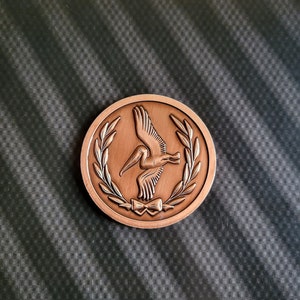 May include: A bronze-coloured coin featuring a pelican in flight, encircled by a laurel wreath. The pelican is facing right and wears a bow tie.