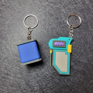 May include: Two keychains with silver rings. One is a blue rectangle with gray trim. The other is a teal, gray, and yellow device with a purple wave display. Both are made of a rubbery material.