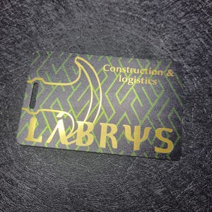 Peut inclure: Une carte rectangulaire avec un motif géométrique vert et des lettres dorées. La carte présente le mot "LABRYS" et le texte "Construction & logistics" en or. La carte a un petit trou en haut.