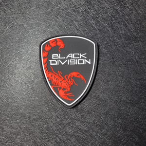 May include: A shield-shaped patch featuring a black background with a red scorpion graphic and the white text "BLACK DIVISION" centered on the surface. The patch has a raised, molded texture and a thin white border outlining the shield shape.