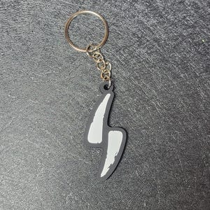 May include: A keychain with a silver ring and chain. The charm is a black lightning bolt shape with white accents. The keychain is on a textured, dark gray surface.