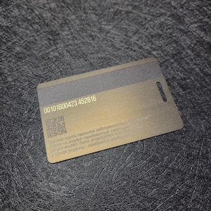 Puede incluir: Una tarjeta rectangular, dorada met&aacute;lica con una franja gris oscuro en la parte superior y un peque&ntilde;o agujero en el borde derecho. La tarjeta tiene una serie de n&uacute;meros y un c&oacute;digo QR impreso. El fondo es una superficie texturizada gris oscuro.