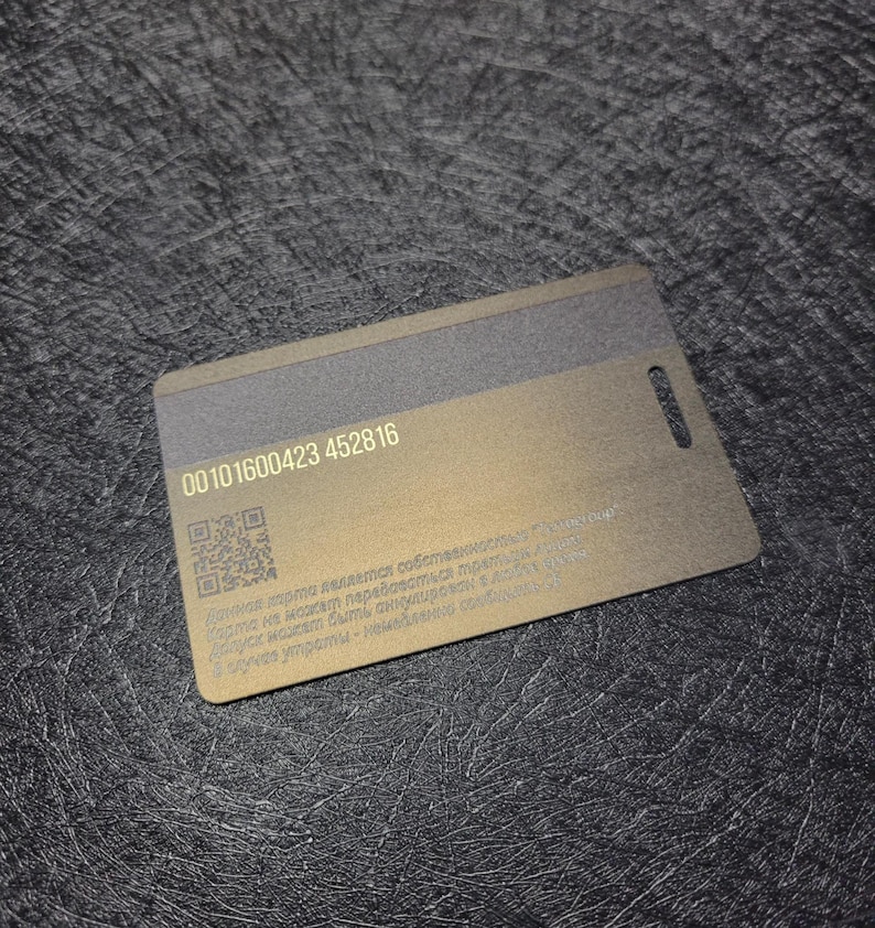 Puede incluir: Una tarjeta rectangular, dorada met&aacute;lica con una franja gris oscuro en la parte superior y un peque&ntilde;o agujero en el borde derecho. La tarjeta tiene una serie de n&uacute;meros y un c&oacute;digo QR impreso. El fondo es una superficie texturizada gris oscuro.