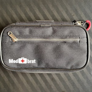 Puede incluir: Bolsa rectangular negra con cremallera plateada y mosquetón rojo. La bolsa tiene el texto "Med brat" en blanco con un símbolo de cruz roja. La bolsa está hecha de un material duradero.