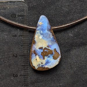Peut inclure: Un pendentif en forme de goutte d'eau avec une pierre d'opale vibrante. L'opale présente un mélange de teintes bleues, blanches et brunes, sur un fond brun foncé. Le pendentif est attaché à un cordon fin brun foncé, créant un collier.