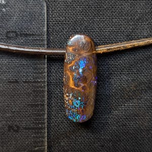 Könnte beinhalten: Ein länglicher, ovaler Anhänger aus dunkelbraunem Boulder-Opal mit irisierenden blauen und grünen Lichtreflexen, befestigt an einer dünnen braunen Kordel. Ein transparentes Lineal links zeigt, dass der Anhänger eine Länge von etwa 3,2 cm aufweist.