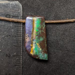 Könnte beinhalten: Ein rechteckiger Anhänger mit polierter Oberfläche, der schillernde grüne, blaue und goldene Flecken auf einer dunkelbraunen Basis aufweist und an einer dünnen Kordel hängt. Ein transparentes Lineal zeigt links eine Länge von 2,5 cm an.
