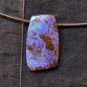 Peut inclure: Un pendentif en opale boulder rectangulaire sur un cordon en cuir marron, présentant des éclats irisés bleus et violets sur une matrice brun foncé. Une règle en plastique transparent est placée à gauche, montrant une longueur d'environ 3,2 cm.