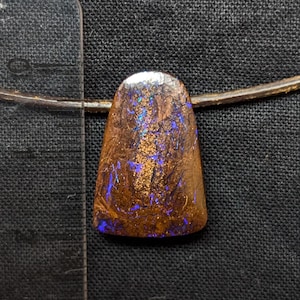 Peut inclure: Un pendentif en opale boulder trapézoïdale présentant une matrice en pierre de fer brun foncé avec des éclats de couleur bleu électrique et or. Le pendentif est suspendu à un cordon fin et placé à côté d'une règle indiquant une longueur de 2,5 cm.