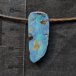 Peut inclure: Un pendentif composé d'une pierre d'opale allongée et irrégulière aux reflets bleus et verts irisés avec des inclusions de matrice brune, suspendu à un cordon brun fin, placé à côté d'une règle transparente indiquant une longueur d'environ 3,8 cm.