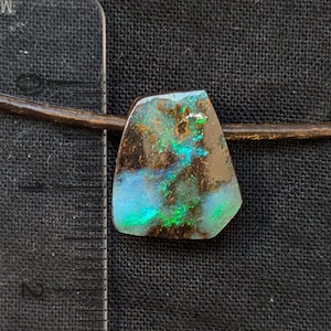 Peut inclure: Un pendentif en opale boulder polie avec des éclats bleus et verts vifs sur un cordon en cuir marron. Une règle en plastique transparent est placée à gauche, montrant que la pierre mesure environ 1,9 cm de largeur au point le plus large.