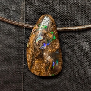 Può includere: Un ciondolo in opale boulder a forma di goccia su un cordino marrone. La pietra presenta una matrice marrone scuro con bagliori iridescenti blu e verdi. Un righello in plastica trasparente a sinistra mostra una lunghezza di circa 3,8 cm.