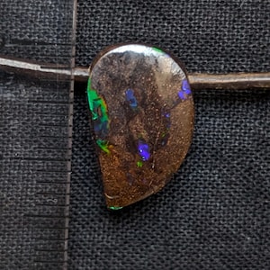 May include: A teardrop-shaped dark brown boulder opal pendant with iridescent green and blue flashes, suspended on a brown cord. A clear plastic ruler is positioned to the left, showing the stone is approximately 1.25 inches in height.
