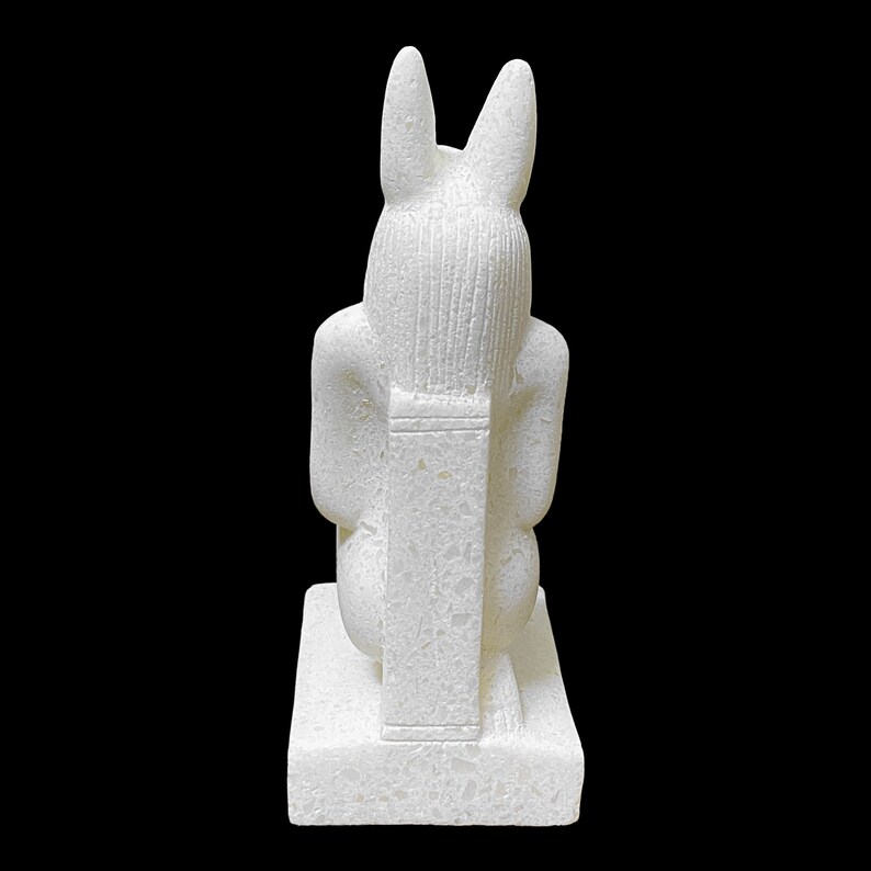 Puede incluir: Estatua de cer&aacute;mica blanca de una deidad egipcia con orejas de conejo, sentada sobre una base.