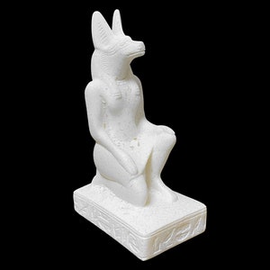Puede incluir: Estatua de cer&aacute;mica blanca de una diosa egipcia con cabeza de chacal, sentada sobre una base con jerogl&iacute;ficos.