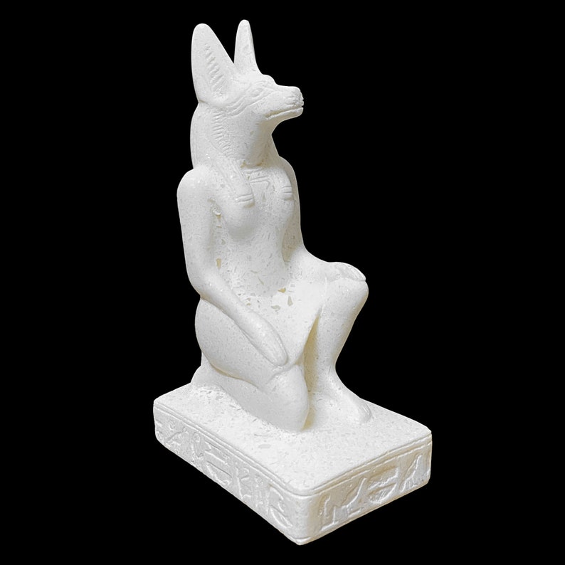 Puede incluir: Estatua de cer&aacute;mica blanca de una diosa egipcia con cabeza de chacal, sentada sobre una base con jerogl&iacute;ficos.