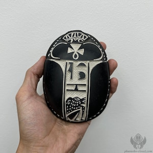 Könnte beinhalten: Schwarzer Skarabäus-Amulett mit weißen geschnitzten Details, darunter Hieroglyphen und ein geflügelter Skarabäus. Das Amulett wird in einer Hand gehalten.