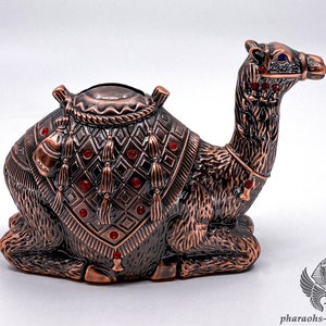 Könnte beinhalten: Eine kupferfarbene Kamelfigur mit roten Juwelen und einem gemusterten Sattel. Das Kamel sitzt mit dem Kopf nach rechts gedreht.