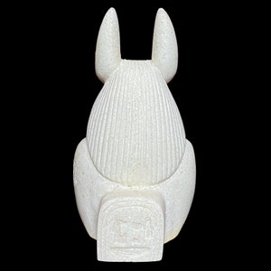 May include: White ceramic statue of the Egyptian goddess Bastet, depicted as a cat with a human head. The statue is sitting with its back to the viewer, and its tail is curled around its legs. The statue has a detailed design, with intricate carvings on its body and head. The statue is about 10 inches tall.