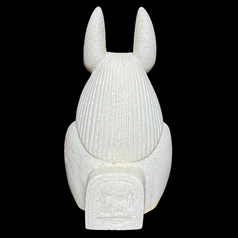 May include: White ceramic statue of the Egyptian goddess Bastet, depicted as a cat with a human head. The statue is sitting with its back to the viewer, and its tail is curled around its legs. The statue has a detailed design, with intricate carvings on its body and head. The statue is about 10 inches tall.