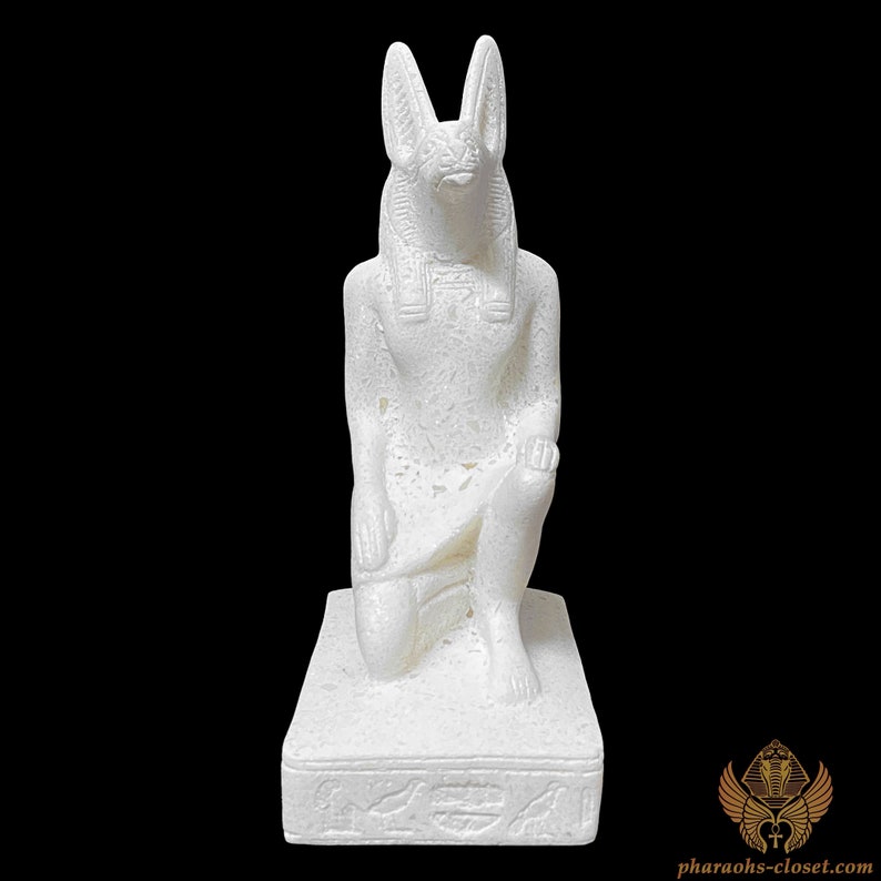 Puede incluir: Estatua blanca de rodillas del dios egipcio Anubis con cabeza de chacal, vistiendo un tocado y una falda tradicionales. La estatua est&aacute; sobre una base rectangular con jerogl&iacute;ficos.