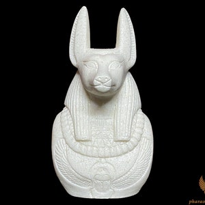 May include: White ceramic statue of Anubis, the jackal-headed Egyptian god of the dead, with a detailed depiction of his head, ears, and body. The statue is sitting upright with its paws folded in front of it.
