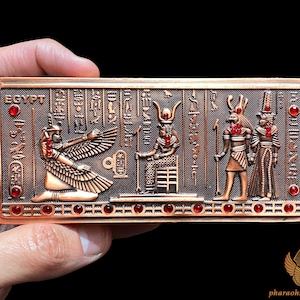 May include: A copper-colored rectangular plaque with an Egyptian hieroglyphic design. The plaque features three figures, including a winged goddess, a seated god, and a standing god. The plaque is decorated with red gemstones.