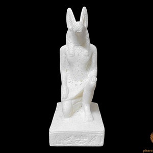 Puede incluir: Estatua de cer&aacute;mica blanca de un dios egipcio con cabeza de chacal, arrodillado sobre una base con jerogl&iacute;ficos. La estatua es detallada y tiene una superficie texturizada.